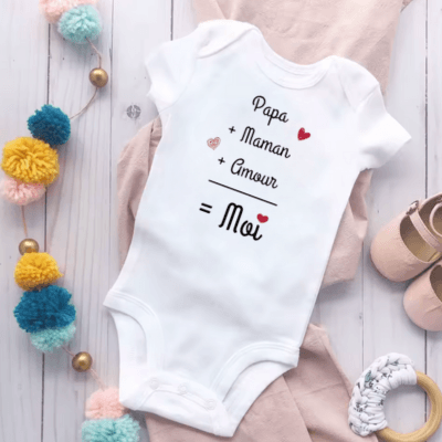 Une borboteuse bebe avec l'inscription - papa plus maman plus amour egal moi -est posée sur un parquet et entourée de petites chaussures et salopette rose et une guirlande de pompons jaunes verts bleus et perles dorées