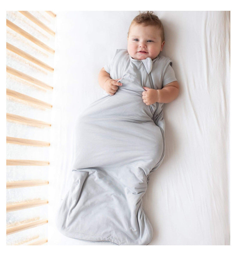 Bébé allongé dans un lit, souriant dans une gigoteuse été grise DouxRefuge™ en bambou, avec un éclairage naturel venant des barreaux du berceau – confort, fraîcheur et bonne humeur pour les nuits chaudes.