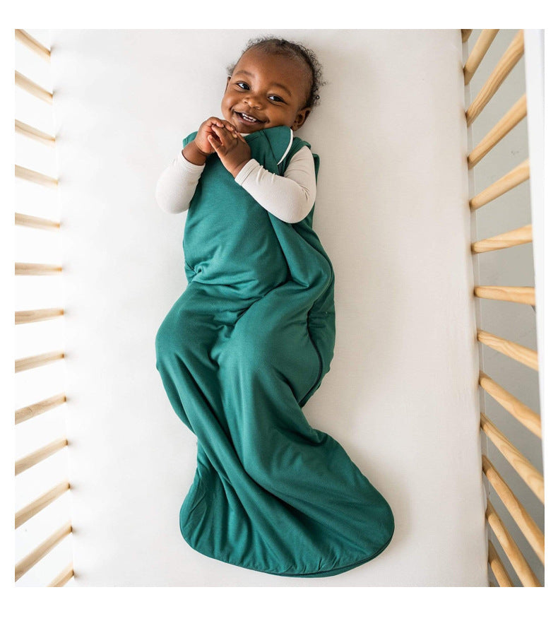 Bébé souriant allongé dans un lit à barreaux, vêtu d’une gigoteuse été DouxRefuge™ en bambou vert foncé – confort, liberté de mouvement et bonne humeur pour les nuits chaudes.