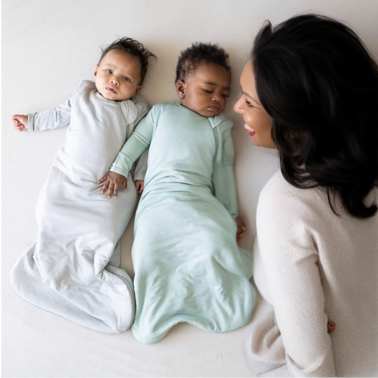Deux bébés en Gigoteuse_Printemps_et_Automne_DouxRefuge™ allongés sur un lit, l’un éveillé et l’autre endormi, accompagnés d’une maman souriante – un moment de douceur et de confort en mi-saison.