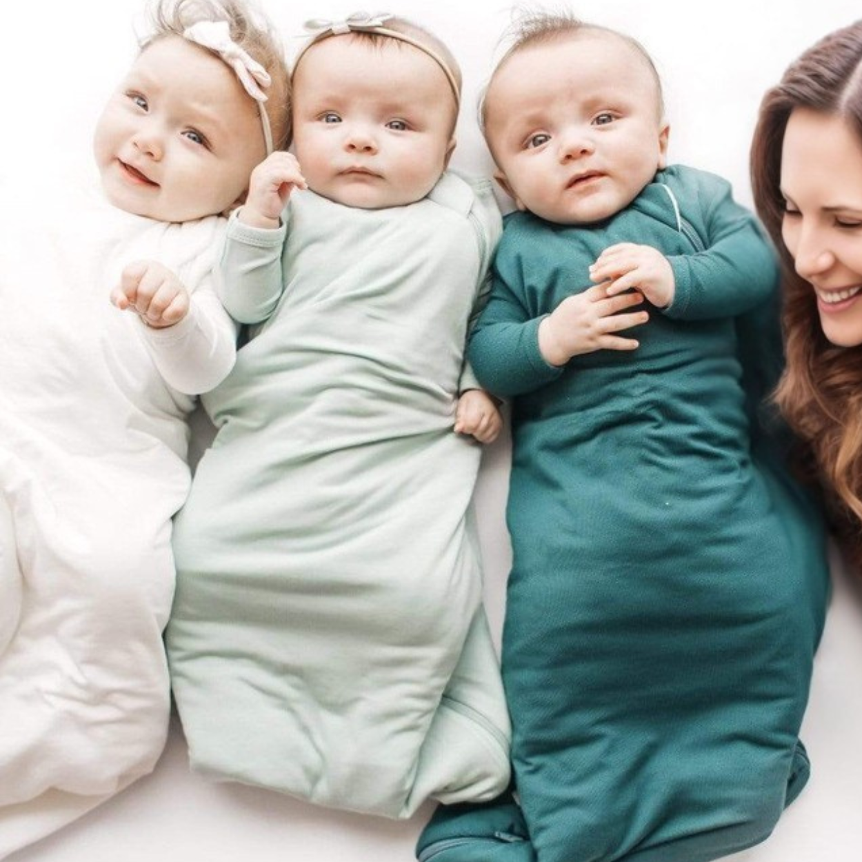Trois bébés allongés côte à côte portant des gigoteuses TOG 0.5 DouxRefuge™ dans les coloris blanc, vert sauge et vert foncé, accompagnés d’une maman souriante – parfaites pour les nuits d’été entre 23 et 26°C.