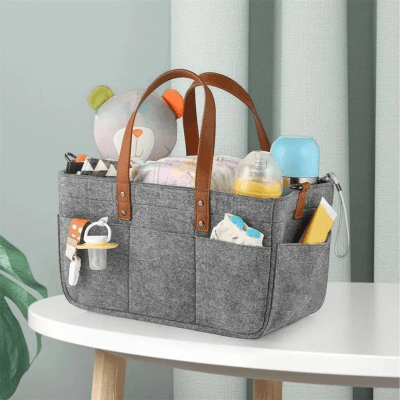 Panier de Rangement Essentiel Bébé™ en feutre gris avec anses en cuir, posé sur une table, rempli de couches, biberon, jouets et accessoires de bébé pour un rangement pratique et organisé