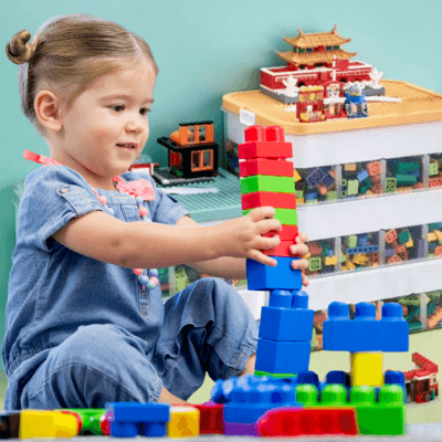 Une petite fille assise qui joue avec des legos bleus rouges vertsnet en arrière plan le Rangement jouet enfant - TidyBox™ empilées avec un couvercle jaune