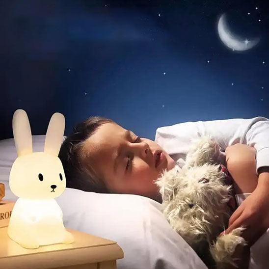 Un petit garçon dort sur le dos dans une chambre bleu avec un ciel étoilé et une lune avec une VEILLEUSE BEBE MonsieurLapinou posé sur la table de chevet