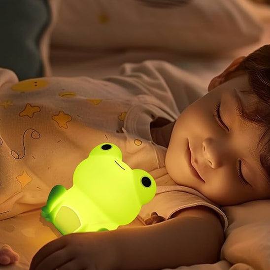 Enfant endormi paisiblement avec une veilleuse bébé grenouille verte allumée posée contre lui, créant une ambiance douce et rassurante pour la nuit."