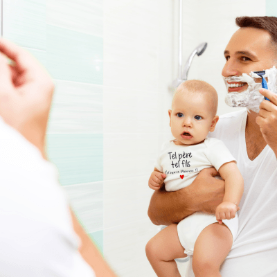 Un père debout devant un miroir se rase en tenant son fils dans un bras et de l'autre se rase. Le bébé porte une barboteuse bebe blanche tel père tel fils
