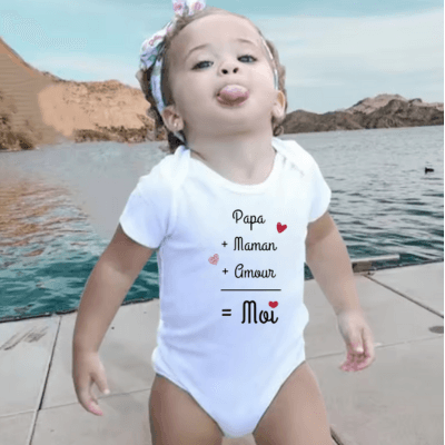 UNE PETITE FILLE QUI TIRE LA LANGUE EST VETUE D'UNE BORBOTEUSE BEBE AVEC L'INSCRIPTION - PAPA PLUS MAMAN PLUS AMOUR EGAL MOI - EST DEBOUT SUR UN PONT AVEC DERIERE ELE LA MER ET UNE BANDE DE TERRE