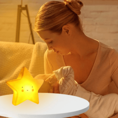 Une veilleuse BEBE VEILLEUSE ETOILE - StarNight™ allumée en jaune et derriere une maman donne le sein à son bebe