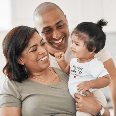 Une maman tient dans ses bras sa petite fille bébé qui porte la barboteuse bebe manche courte blanche tel père telle fille et derière se tient son mari. Tous les trois ont un large sourire 