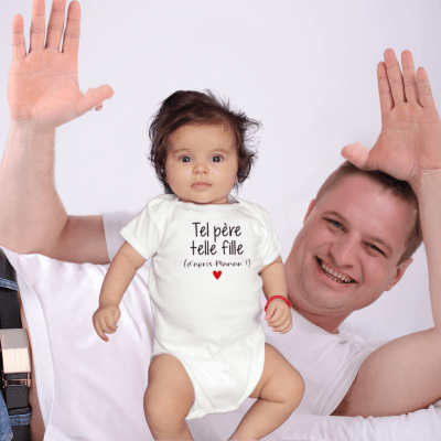 Un père allongé sur le côté les deux bras en l'air sa petite fille bébé debout devant lui porte la barboteuse bebe manche courte blanche tel peère telle fille