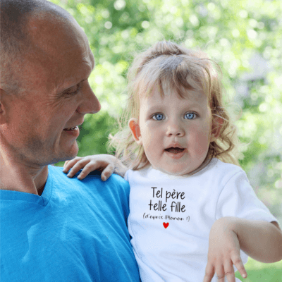 Un père tient dans ses bras sa petite fille bébé blonde aux yeux bleus qui porte la barboteuse bebe manche courte blanche tel père telle fille