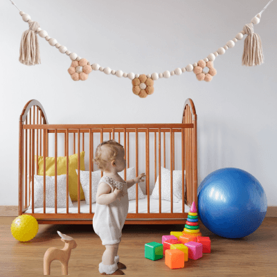 GUIRLANDE - PomponsBoho accroche au mur dans une chambre de bebe couleurs claires avec un bebe debout devant le berceau
