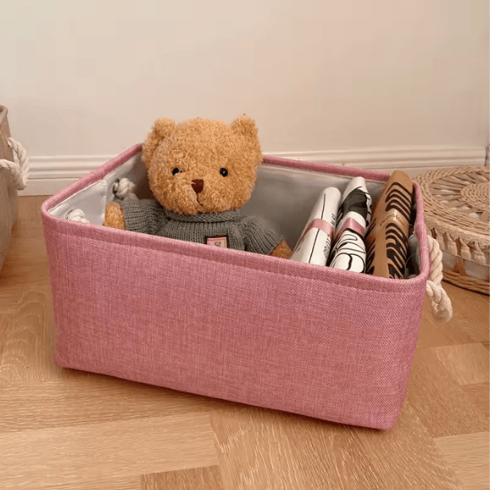 Panier de Rangement Pour Bébé - Rose posée au sol avec un ours en peluche dedans et des T-shirt 