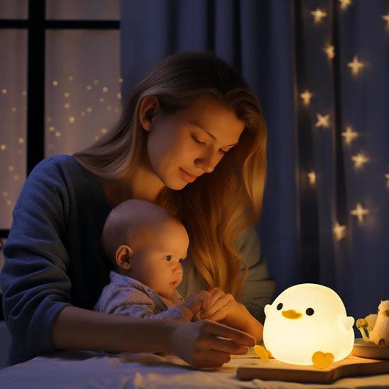 Un maman assise à une table à la lumière d'une VEILLEUSE BEBE CANETON lit son bébé sur ses ses genoux 