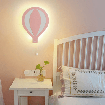 Une Veilleuse bébé Murale Marco Polo rose sur un mur rose pale au dessus d'une table de chevet blanche sur laquel est posée une plante verte dans un vase en verre et à droite une tête de lit à bareaux blanc un coussin beige posé contre la tête de lit