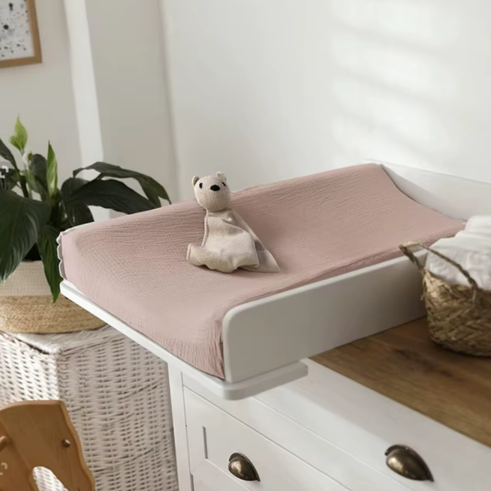 Housse pour matelas à langer rose installée sur une table à langer, avec un petit doudou posé dessus dans une chambre de bébé.