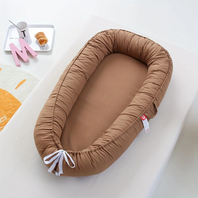 Réducteur de lit bébé marron MokaCocon™ avec ruban blanc, présenté sur une table dans une ambiance chaleureuse.